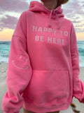 Person at the beach wearing a pretty pink oversized hoodie with 'HAPPY TO BE HERE' text.