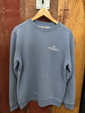 A blue crewneck sweatshirt with a small embroidered text on the chest, displayed on a hanger against a wooden background.
