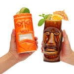 Two tiki-themed glasses held by hands, one orange and one brown, with drinks and garnishes.